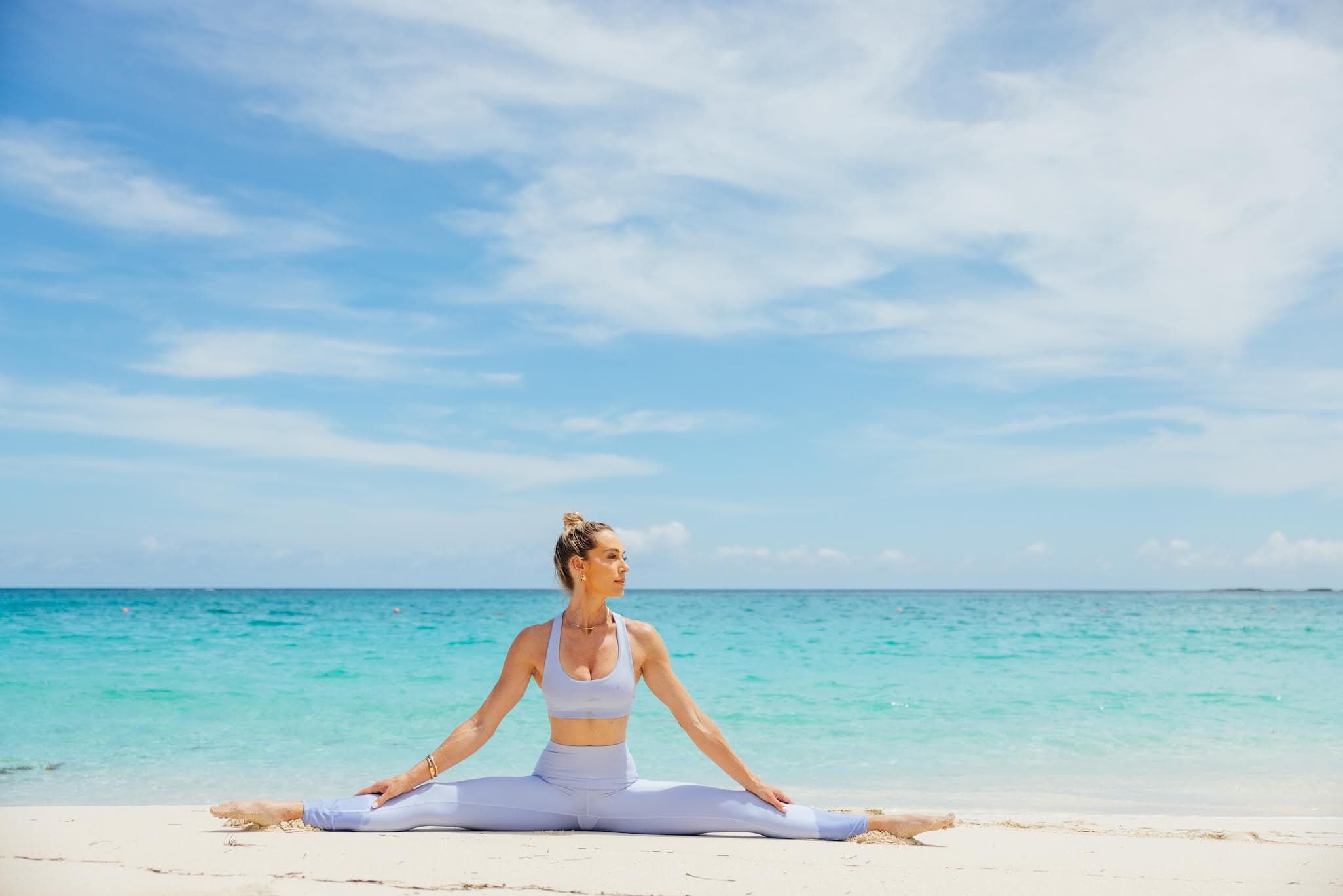 Nora seated in a wide-legged stretch on white sand beside turquoise water